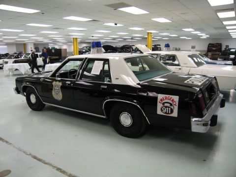 1991 Ford LTD Crown Victoria - black-over-white police cruiser with a gold door shield, blue beacon and Emergency 911 decal parked in an indoor collection.