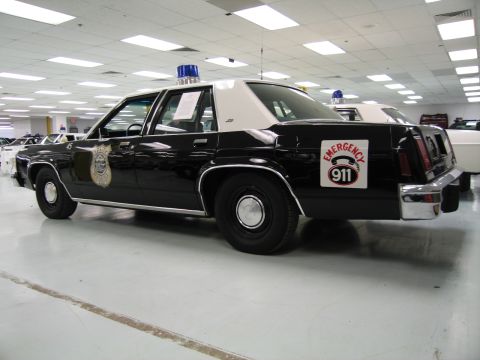 1991 Ford LTD Crown Victoria - Rear three-quarter view of a black-over-white police sedan showing a door shield, blue roof light and Emergency 911 decal.
