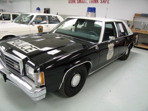 1991 Ford LTD Crown Victoria - black-over-white police sedan marked POLICE on the hood with car number 45 and a blue light bar, parked in a workshop garage.