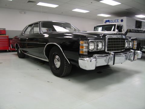 1991 Ford LTD Crown Victoria - black 1970s full-size Ford four-door sedan with stacked headlights, parked indoors near a marked police van.