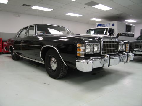 1991 Ford LTD Crown Victoria - black 1970s Ford four-door with chrome push-bar bumper guards displayed beside a police van inside a garage.