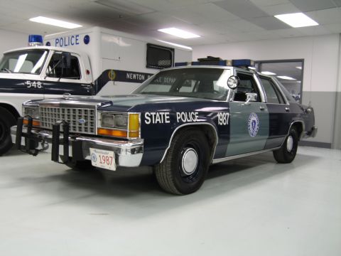1991 Ford LTD Crown Victoria - blue State Police Ford sedan lettered 1987 with a state seal on the door, parked beside a marked police van indoors.