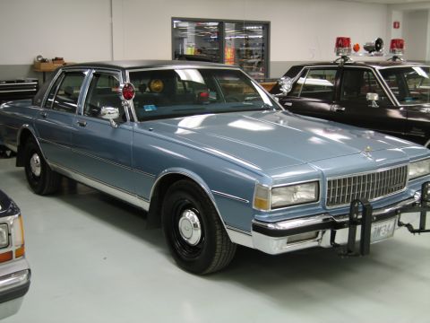 1991 Ford LTD Crown Victoria - light-blue 1980s Chevrolet Caprice four-door fitted with a pillar spotlight and front push bar, parked in a collection garage.