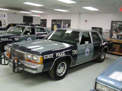 1991 Ford LTD Crown Victoria - two-tone green State Police Ford sedan numbered 700 with a door seal, displayed among other cruisers in an indoor hall.