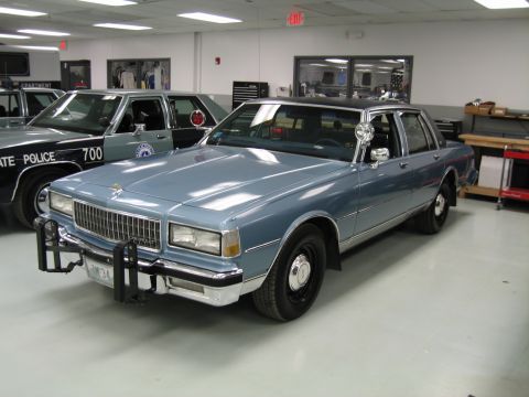 1991 Ford LTD Crown Victoria - light-blue 1980s Chevrolet Caprice four-door with a spotlight and front push bar, parked beside a green State Police car.