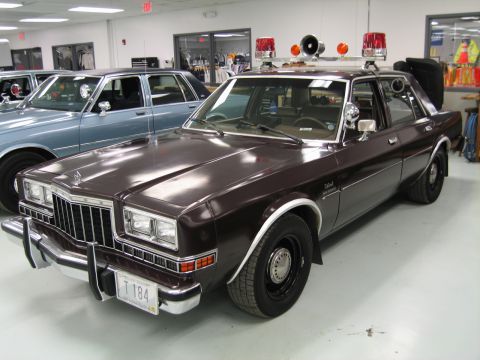 1991 Ford LTD Crown Victoria - maroon-brown 1980s four-door sedan fitted with twin pillar spotlights and a roof rack of red emergency lights, parked indoors.