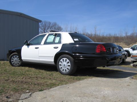 1991 Ford LTD Crown Victoria - Rear three-quarter view of a black-and-white Ford Crown Victoria police sedan parked on grass beside a metal building.