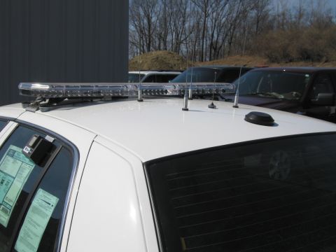 1991 Ford LTD Crown Victoria - Close-up of a white police car's roof showing a long chrome light bar, antennas and window paperwork, photographed outdoors.