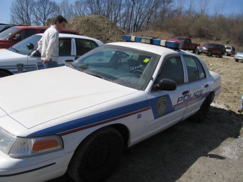 1991 Ford LTD Crown Victoria - white police sedan marked POLICE with a blue stripe and roof lightbar sits on a gravel lot as a man stands at the next car.