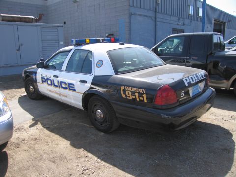 1991 Ford LTD Crown Victoria - Rear three-quarter view of a black-and-white Billerica Police sedan with a roof lightbar and EMERGENCY 9-1-1 markings in a dirt lot.