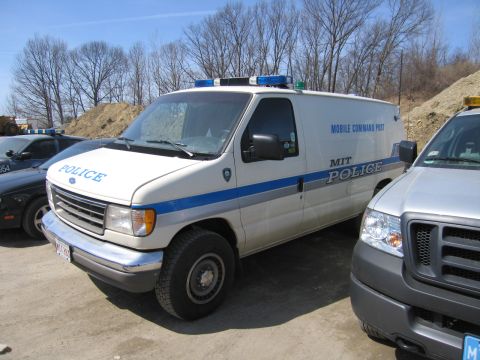 1991 Ford LTD Crown Victoria - white Ford full-size van lettered MIT Police Mobile Command Post with a roof lightbar sits among other vehicles on a gravel lot.