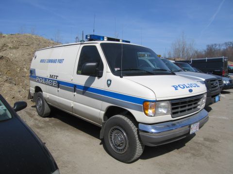 1991 Ford LTD Crown Victoria - Front three-quarter view of a white Ford MIT Police Mobile Command Post van with a roof lightbar parked on a dirt lot under blue sky.