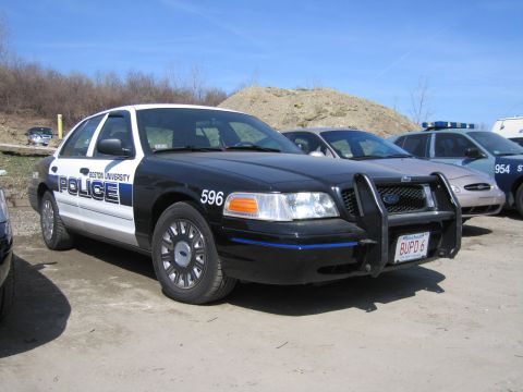 1991 Ford LTD Crown Victoria - black-and-white Boston University Police sedan numbered 596 with a front push bar and BUPD 6 plate parked on a dirt lot.