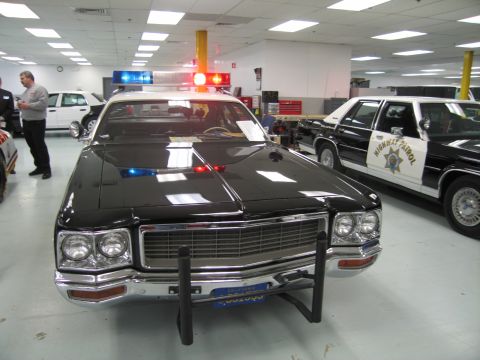1991 Ford LTD Crown Victoria - Head-on view of a black vintage full-size police sedan with chrome grille, roof emergency lights and push bar among display cars indoors.