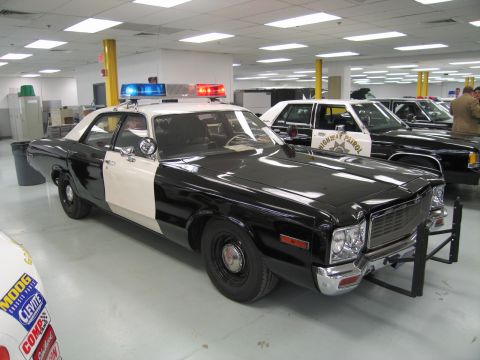 1991 Ford LTD Crown Victoria - black-and-white vintage two-door police car with roof lights and a front push bar displayed on a showroom floor near other cruisers.