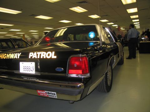 1991 Ford LTD Crown Victoria - Rear view of a black Ford Highway Patrol sedan with a lit roof beacon and plate 888849 displayed indoors as people stand nearby.