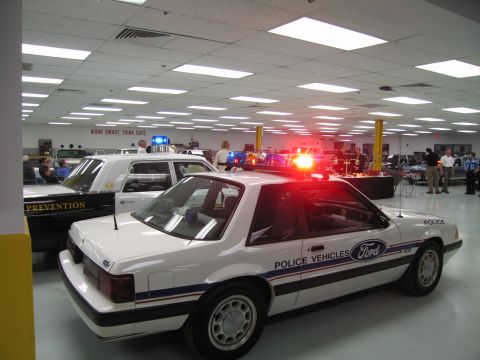 1991 Ford LTD Crown Victoria - white Ford Mustang police car lettered Ford Police Vehicles with lit roof lights sits among other display cruisers in a showroom.
