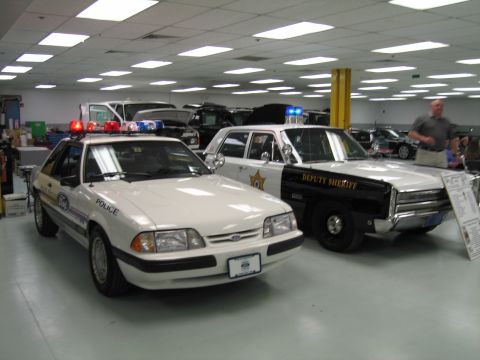 1991 Ford LTD Crown Victoria - white Ford Mustang police car parked beside a black-and-white Deputy Sheriff cruiser in an indoor showroom with other vehicles behind.