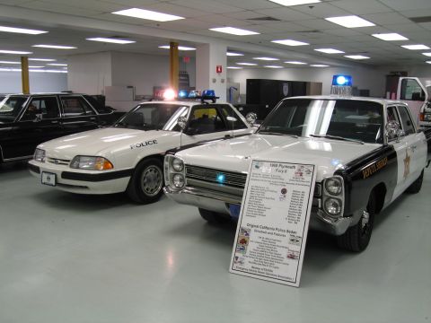 1991 Ford LTD Crown Victoria - white Ford Mustang police car next to a black-and-white vintage Plymouth police sedan with an info placard in an indoor display.