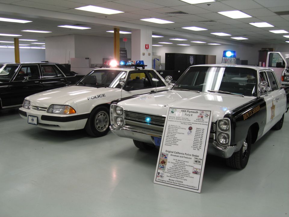 White Ford Mustang police car beside black and white vintage Plymouth sedan indoors