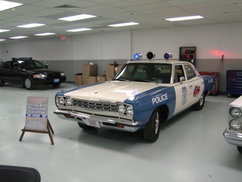 1991 Ford LTD Crown Victoria - blue-and-white 1968 Plymouth Satellite police sedan with roof lights and a descriptive easel placard displayed on a showroom floor.