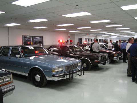 1991 Ford LTD Crown Victoria - row of 1970s and 1980s police sedans, led by a blue Chevrolet Caprice with push bar, parked indoors as men converse beside them.