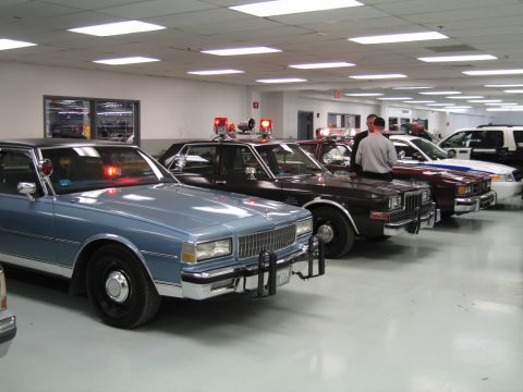 1991 Ford LTD Crown Victoria - row of vintage police sedans led by a blue Chevrolet Caprice with roof lights and push bar parked on an indoor showroom floor.