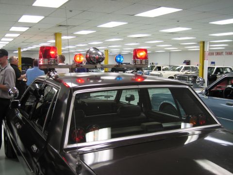 1991 Ford LTD Crown Victoria - Close view of a vintage police car's roof-mounted red and blue dome lights and chrome pillar spotlight inside an indoor showroom.