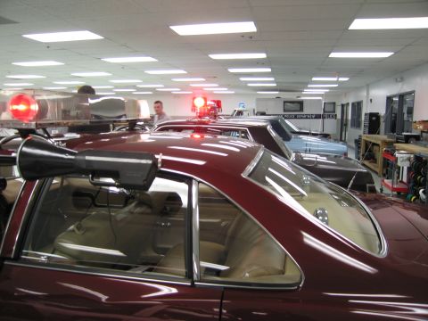 1991 Ford LTD Crown Victoria - Maroon sedan roofline fitted with a rotating red beacon and a roof-mounted loudspeaker, photographed inside a display hall.