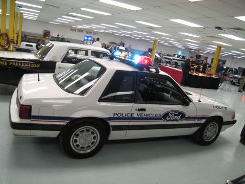 1991 Ford LTD Crown Victoria - White Ford Mustang fox-body police car with lit lightbar and Police Vehicles door graphics, shown in an indoor display.