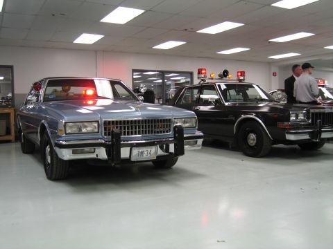 1991 Ford LTD Crown Victoria - Silver-blue Chevrolet Caprice police car with push bar and lit reds parked beside a black patrol sedan in a showroom.