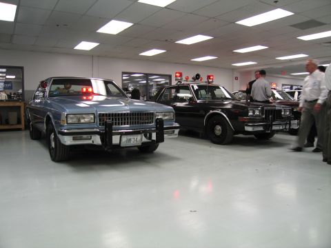 1991 Ford LTD Crown Victoria - Silver Chevrolet Caprice and a black patrol sedan with red lights on, surrounded by visitors in an indoor display hall.