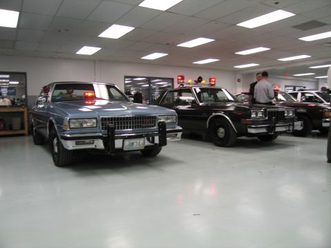 1991 Ford LTD Crown Victoria - Silver Chevrolet Caprice police car with push bar beside a black patrol sedan, both with lights on inside a display hall.