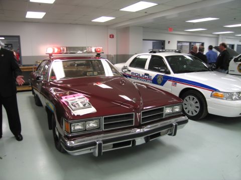 1991 Ford LTD Crown Victoria - Maroon vintage patrol sedan with a lit lightbar next to a white Ford Crown Victoria Police Interceptor in an indoor display.