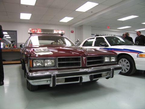 1991 Ford LTD Crown Victoria - Front view of a maroon vintage patrol sedan with quad headlights beside a white Crown Victoria police car in a showroom.