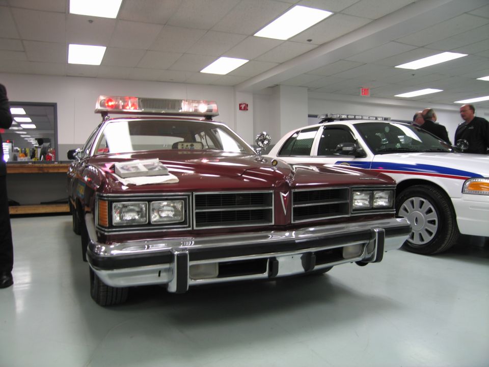 Front of maroon vintage sedan beside white Crown Victoria police car indoors