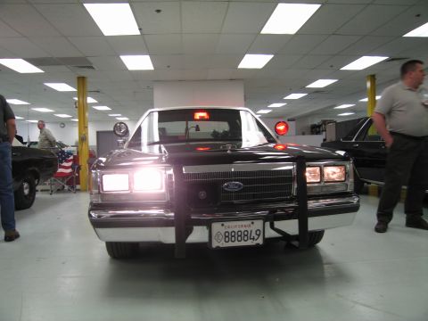 1991 Ford LTD Crown Victoria - Black Ford patrol sedan with chrome push bar and California plate 888849, lit headlights, with people standing alongside indoors.