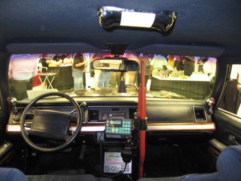 1991 Ford LTD Crown Victoria - Police car cabin showing the steering wheel, mounted radio equipment and a red vertical shotgun lock, viewed from the rear seat.