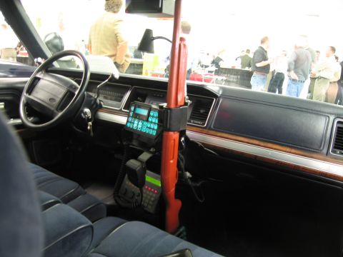 1991 Ford LTD Crown Victoria - Police car cabin showing the steering wheel, mounted radio keypad and microphone, and a red vertical shotgun mount.