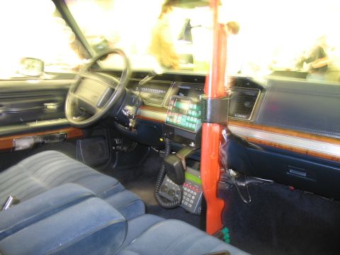 1991 Ford LTD Crown Victoria - Police car interior with blue bench seat, mounted radio keypad and microphone, and an orange vertical shotgun mount.