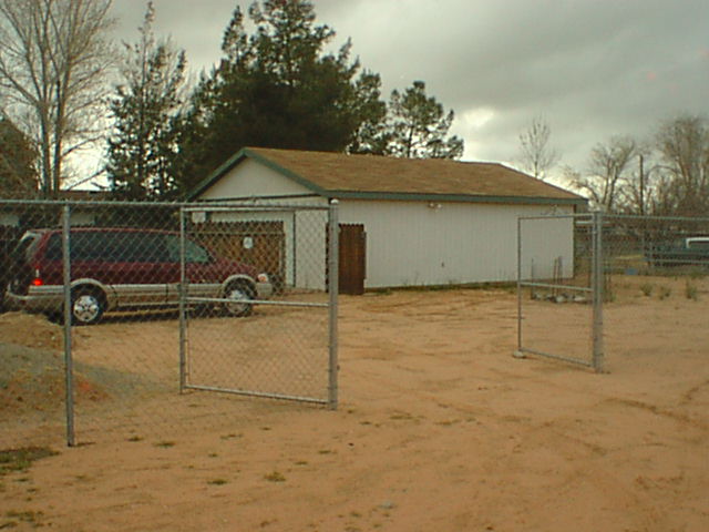 Red SUV parked beside a tan metal outbuilding inside a chain-link fenced dirt lot