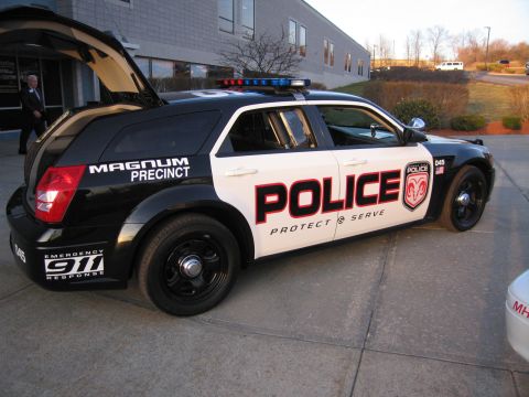 1991 Ford LTD Crown Victoria - Passenger-side view of a black and white police Dodge Magnum wagon lettered POLICE Protect & Serve, with open liftgate and roof light bar.