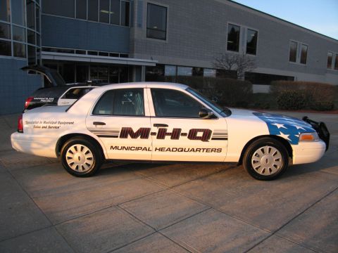 1991 Ford LTD Crown Victoria - Side view of a white Ford Crown Victoria sedan with M-H-Q Municipal Headquarters lettering and flag graphics, parked outside a building.