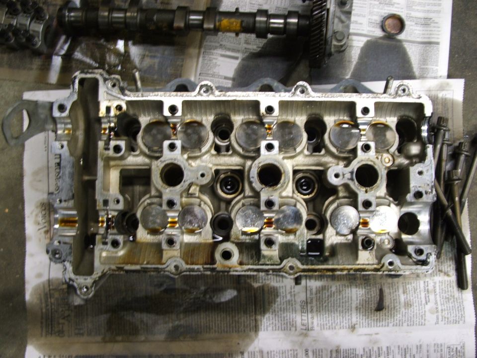 Disassembled engine cylinder head with camshaft and valves visible, resting on newspaper