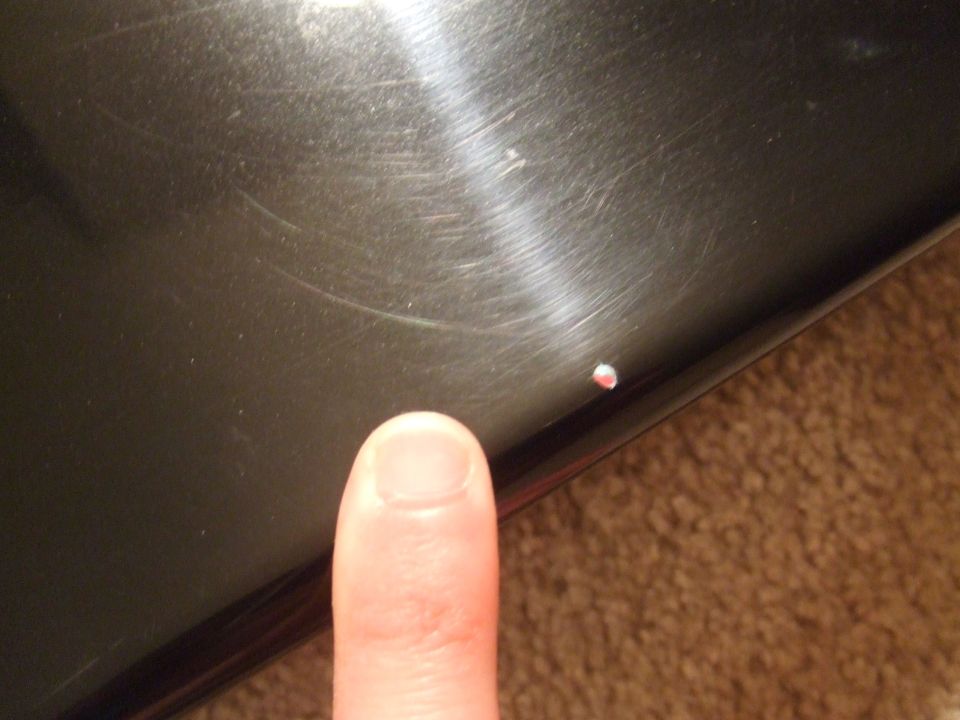 Finger pointing at a small chip on a dark, possibly metallic surface. Carpet visible in background