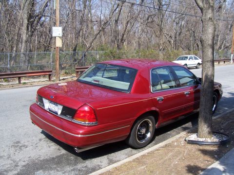 1999 Ford Crown Victoria - A red sedan is parked on the side of a street next to a tree, with another white car visible in the background.