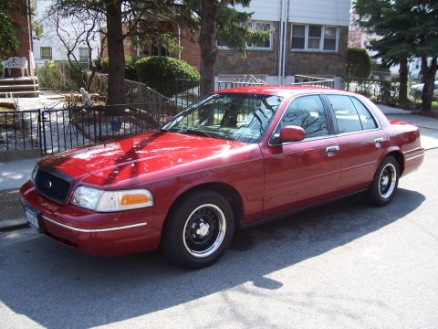1999 Ford Crown Victoria - A red sedan is parked on a residential street with a house and trees in the background.