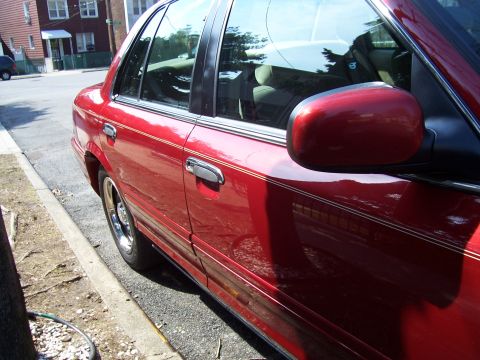 1999 Ford Crown Victoria - The side of a red sedan is visible, parked along a curb on a sunny day.