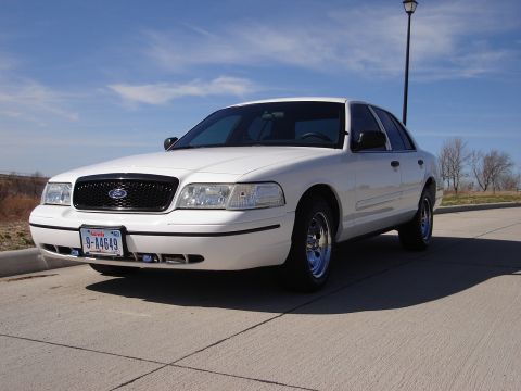 2000 Ford Crown Victoria - This white sedan is parked on a paved surface under a clear blue sky with a few trees in the background.