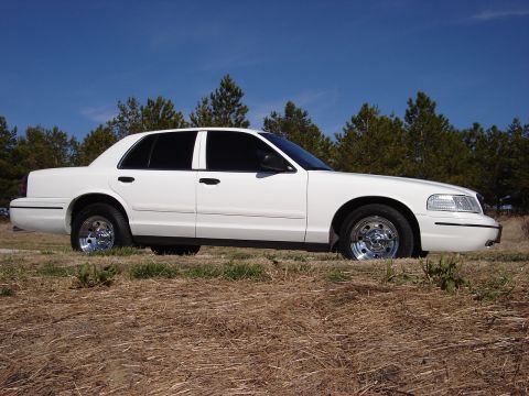 2000 Ford Crown Victoria - A white 2000 Ford Crown Victoria sedan is viewed from the side, parked on a dry, grassy field with trees in the background.